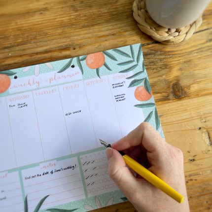 A weekly planner with an illustrated design featuring oranges and leaves sits on a table. Above it, a woman holds a yellow fountain pen. A cup of tea sits off to the side.