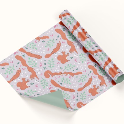 A roll of wrapping paper sits half unrolled on a white background. The wrapping paper depicts a pattern featuring red squirrels surrounded by hearts, arrows, clover, leaves and baby's breath flowers. The pattern has a lilac background.