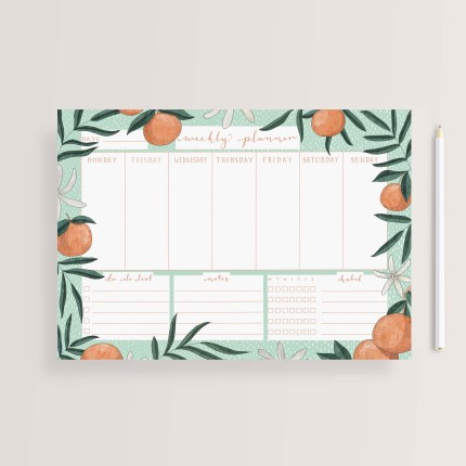 A weekly planner with a pen next to it features a fun pattern of oranges and leaves against a light green background.