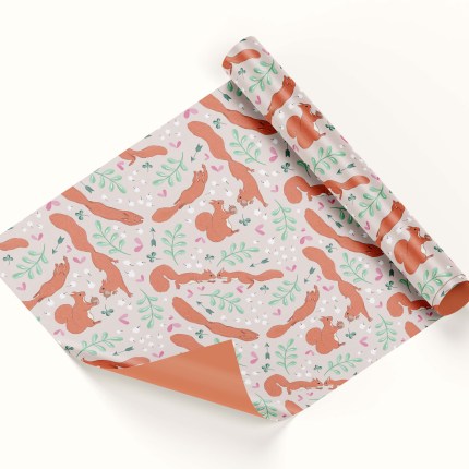 A roll of wrapping paper sits half unrolled on a white background. The pattern features red squirrels, green clover, leaves and arrows, with white baby's breath flowers, on a light pink background.